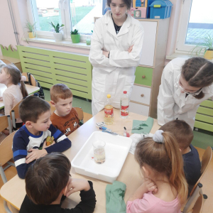 Przedszkolaki siedzą przy stole. Na środku biała miska ze szklaną wlewką. Uczestniczki kółka chemicznego przeprowadzają eksperyment chemiczny dla dzieci.  Kliknięcie na zdjęcie spowoduje jego powiększenie do rozmiaru oryginalnego.