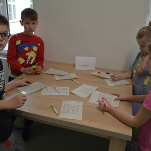 Dzieci stoją przy stoliku. Rozwiązują sudoku, które leży przed nimi.  Kliknięcie na zdjęcie spowoduje jego powiększenie do rozmiaru oryginalnego.