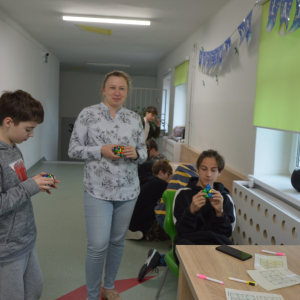 Pani Justyna Solak trzyma w rękach kostkę Rubika. Obok jeden z chłopców układa kostkę Rubika towarzyszy mu drugi uczeń, który również bawi się kostką.  Kliknięcie na zdjęcie spowoduje jego powiększenie do rozmiaru oryginalnego.